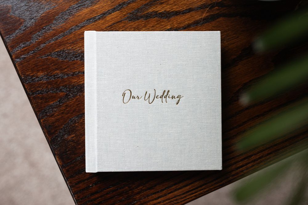 A minimalist white wedding album with gold lettering on a dark wooden table.