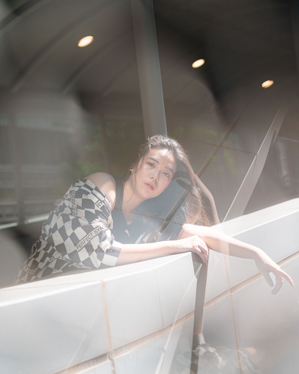 Artistic portrait through glass with moody lighting and a checkered jacket creating reflective effects.