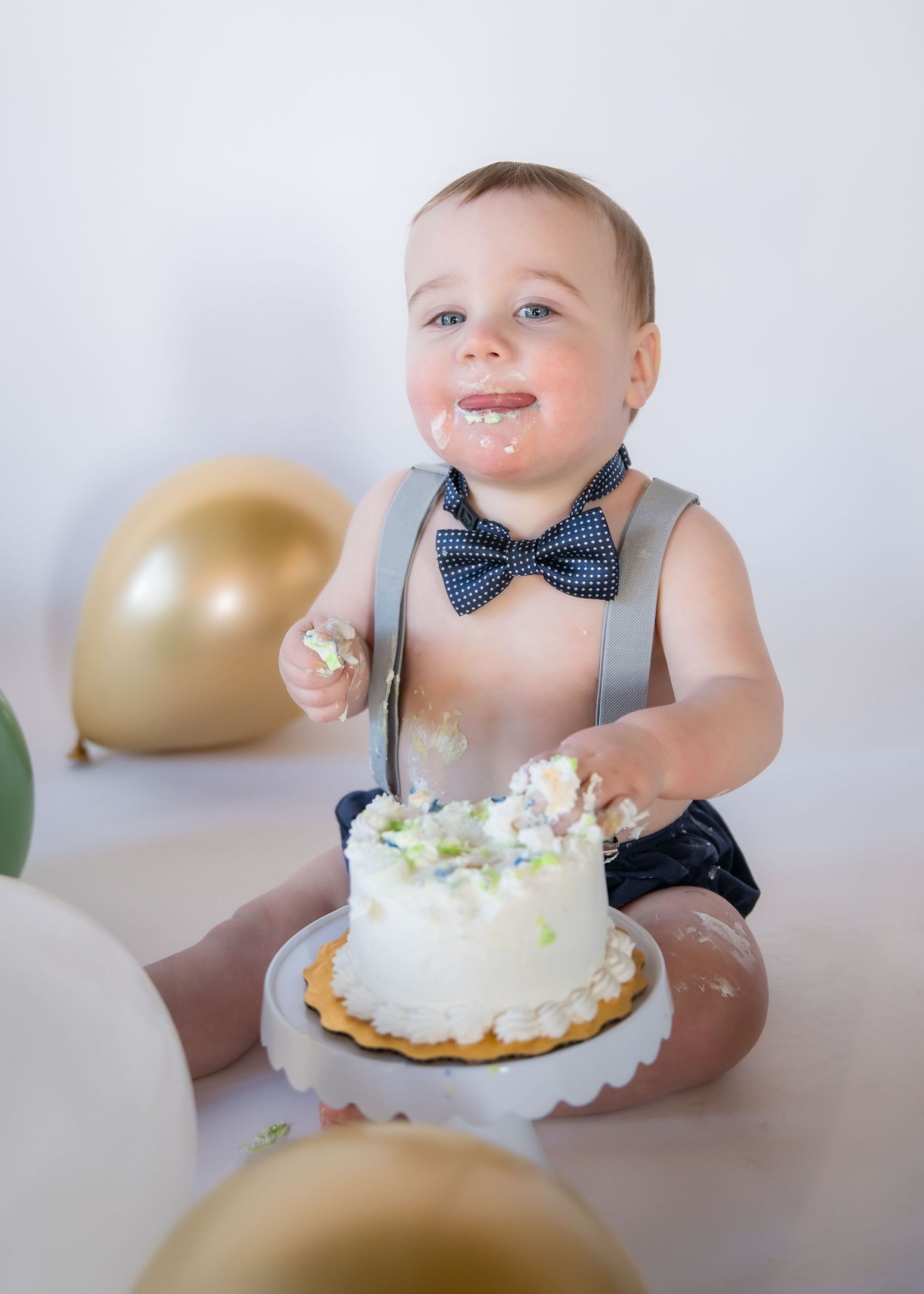 Baby wearing suspenders and bow tie enjoys first birthday cake with gold and white balloons in background.