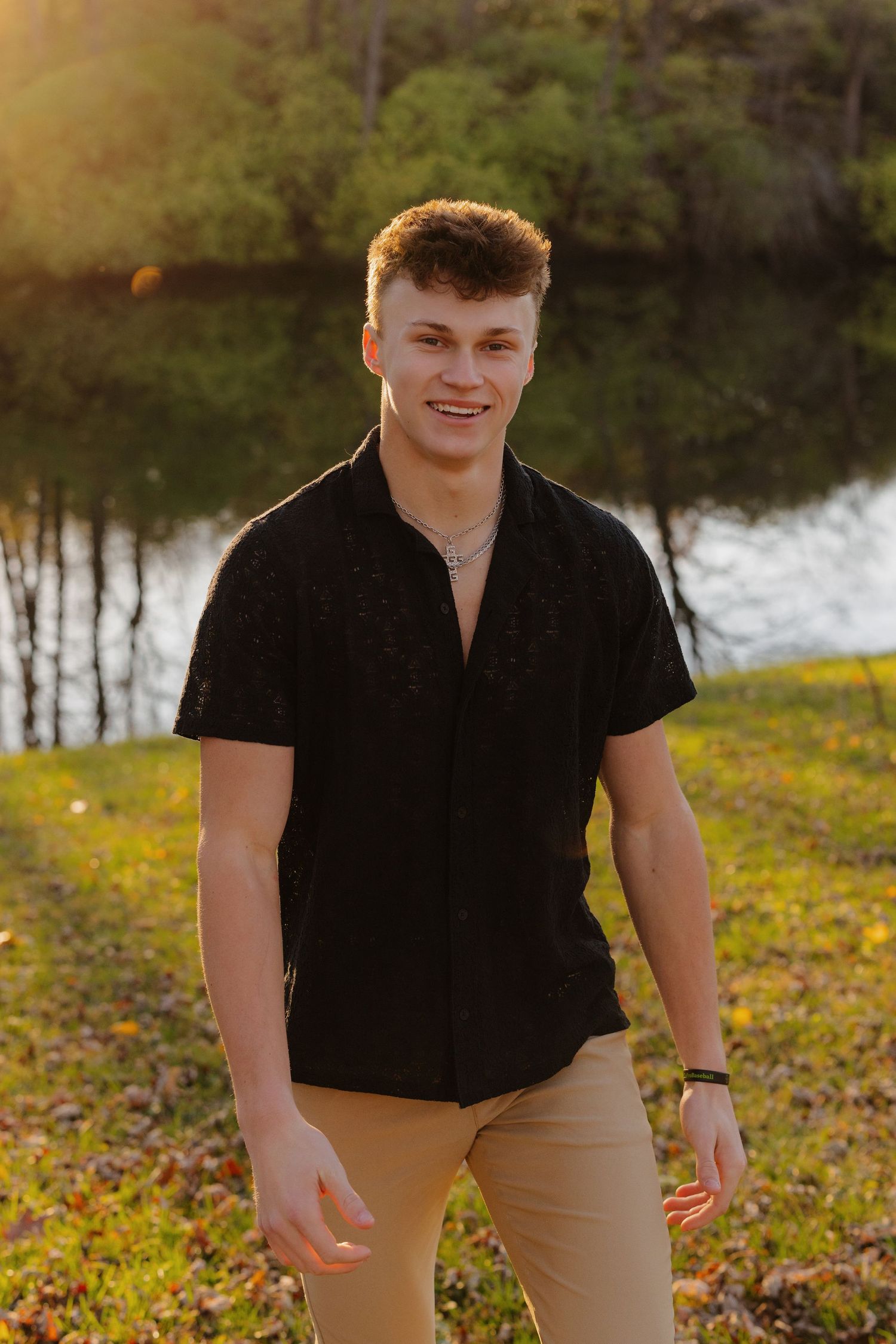 Young person in black polo shirt and khakis poses outdoors by a lake during golden hour sunset.
