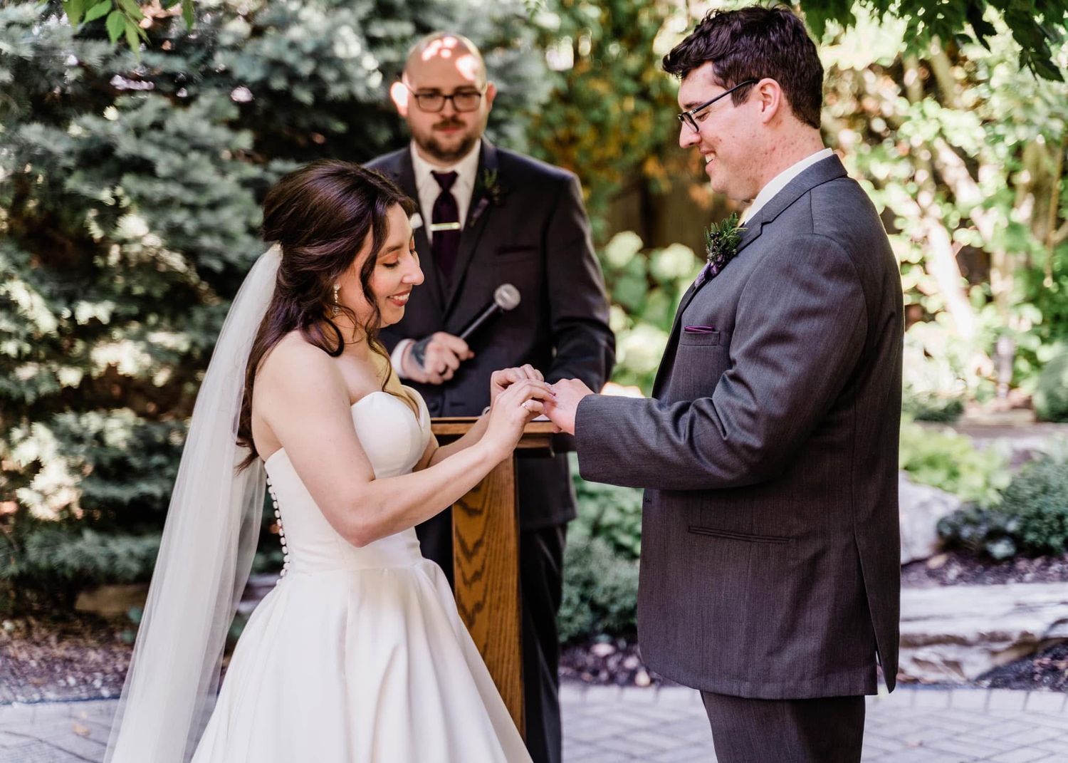 A couple exchanges rings during an outdoor wedding ceremony officiated by a minister in a garden setting.