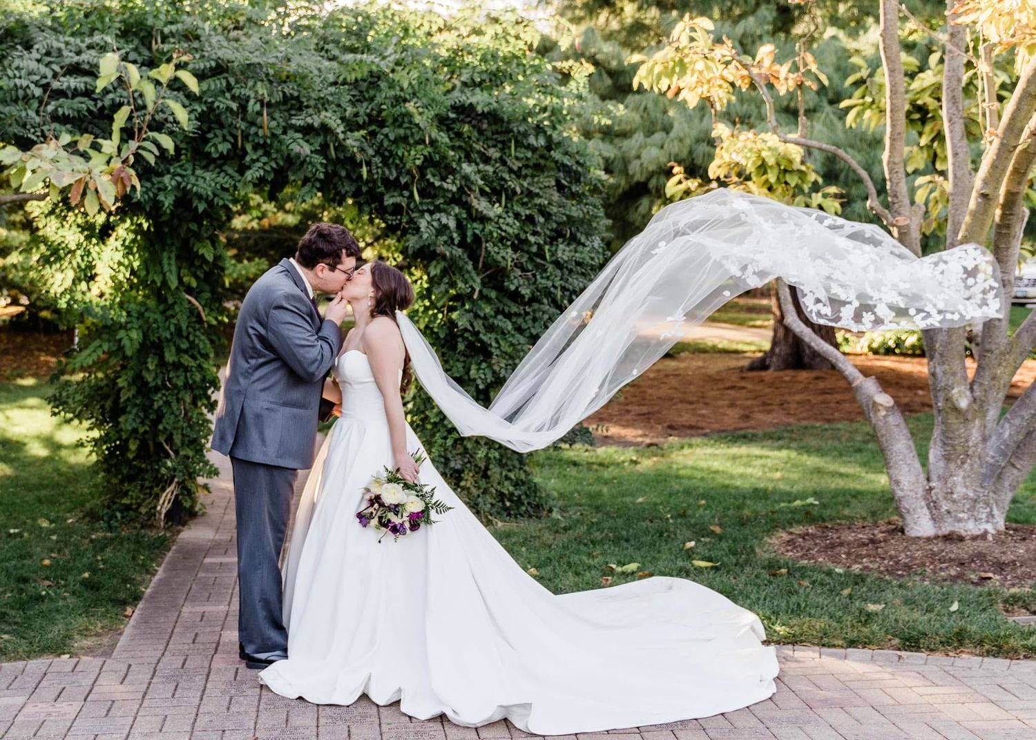 A romantic garden wedding moment as the bride's long veil flows in the breeze on a landscaped wooden pathway.