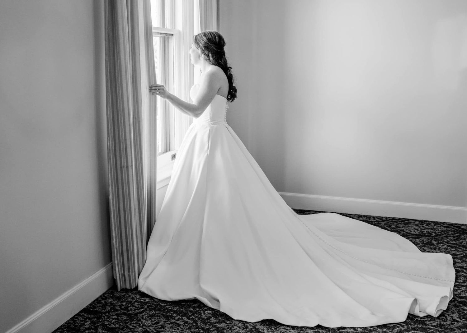 A bride in an elegant white ball gown with sweeping train stands looking out a window in classic black and white.