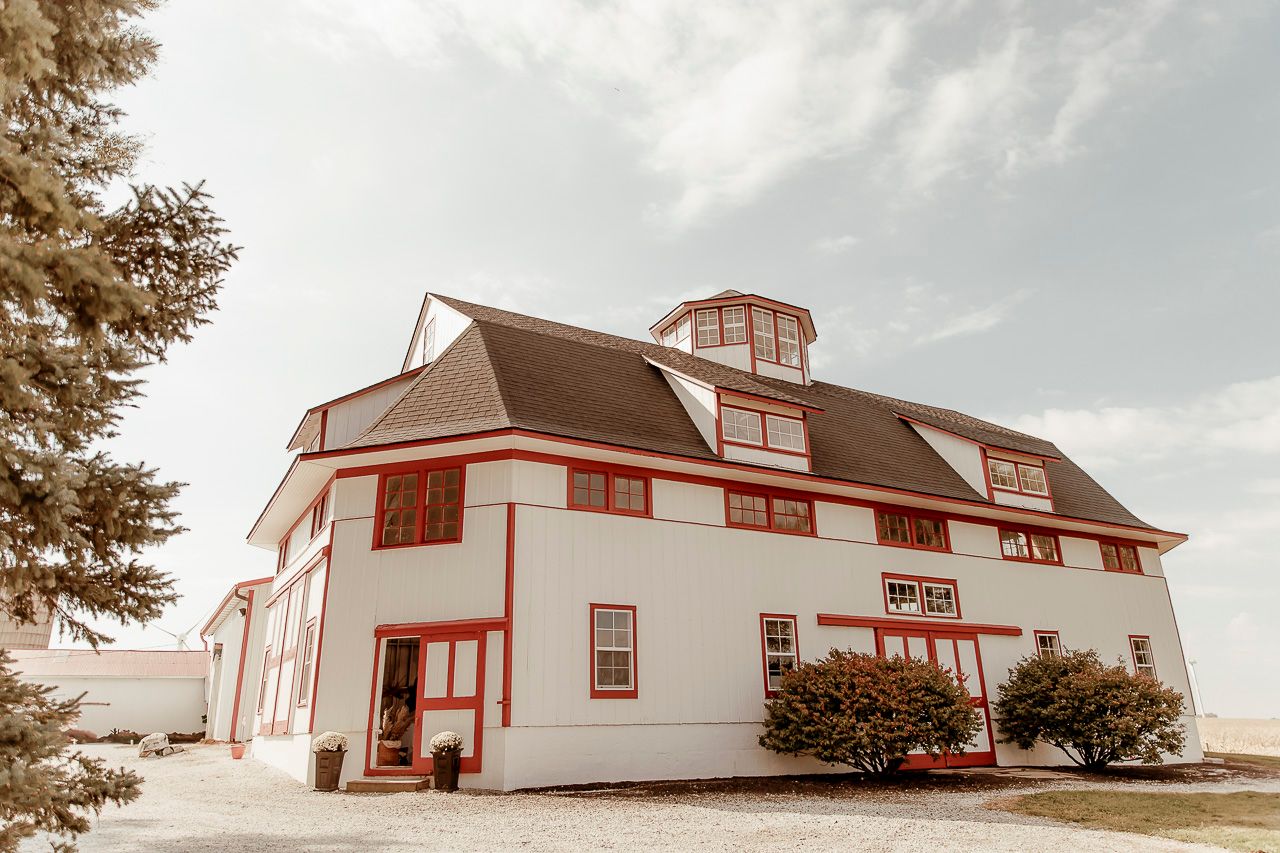 A white barn-style building with red trim and a cupola sits against a cloudy sky in a rustic countryside setting.