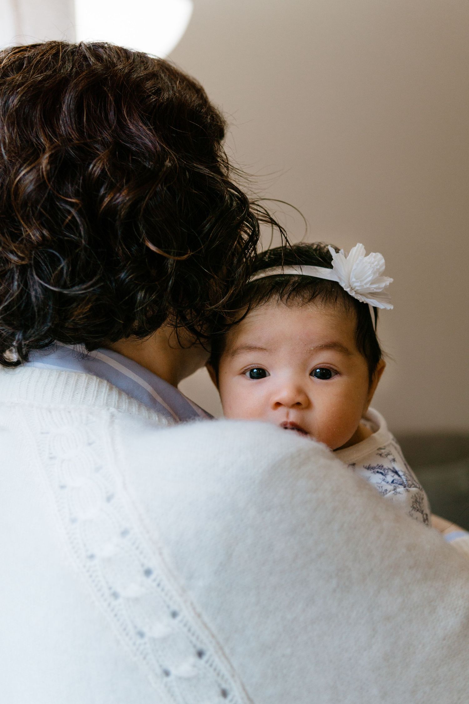 Baby wearing a white headband with flower accessory peeks over grandmother's shoulder.