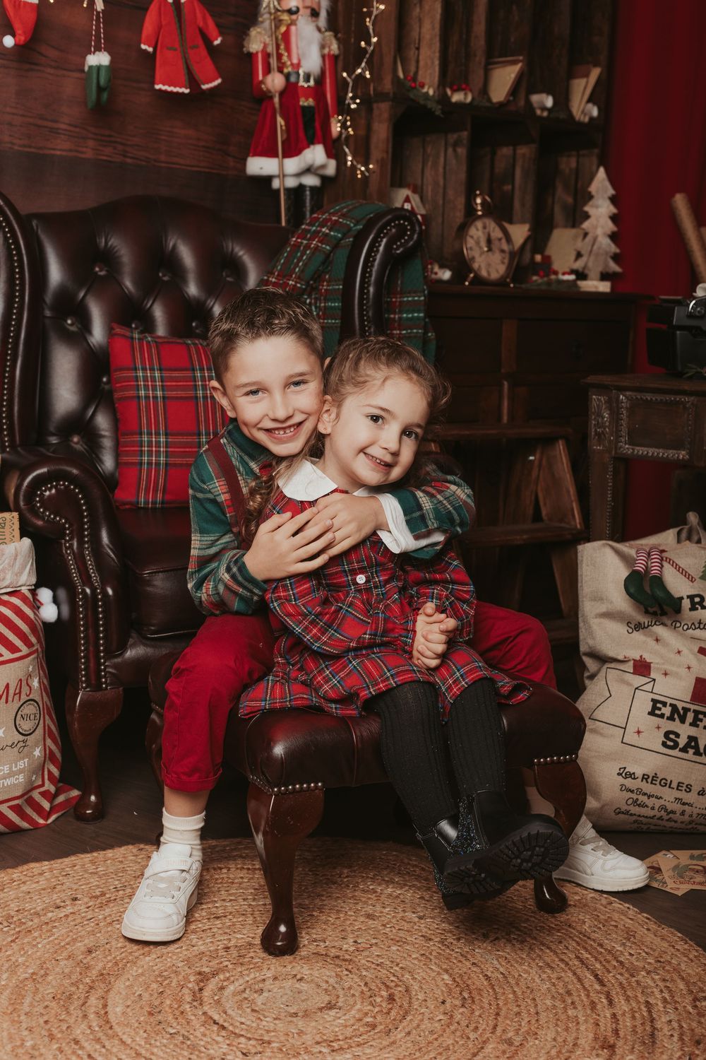 A festive holiday scene with two children sitting in a leather armchair surrounded by Christmas decorations and plaid accents.