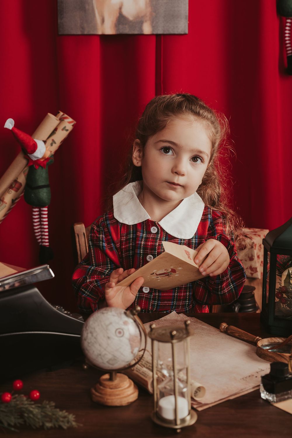 Child in plaid dress writes letter to Santa at wooden desk with vintage lantern and holiday decorations.