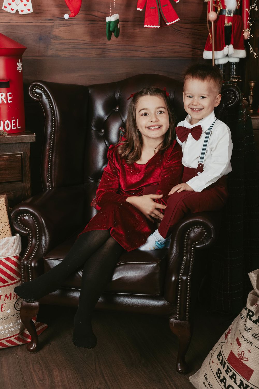 Two children in festive holiday attire sit together in a dark leather armchair with Christmas decorations in the background.
