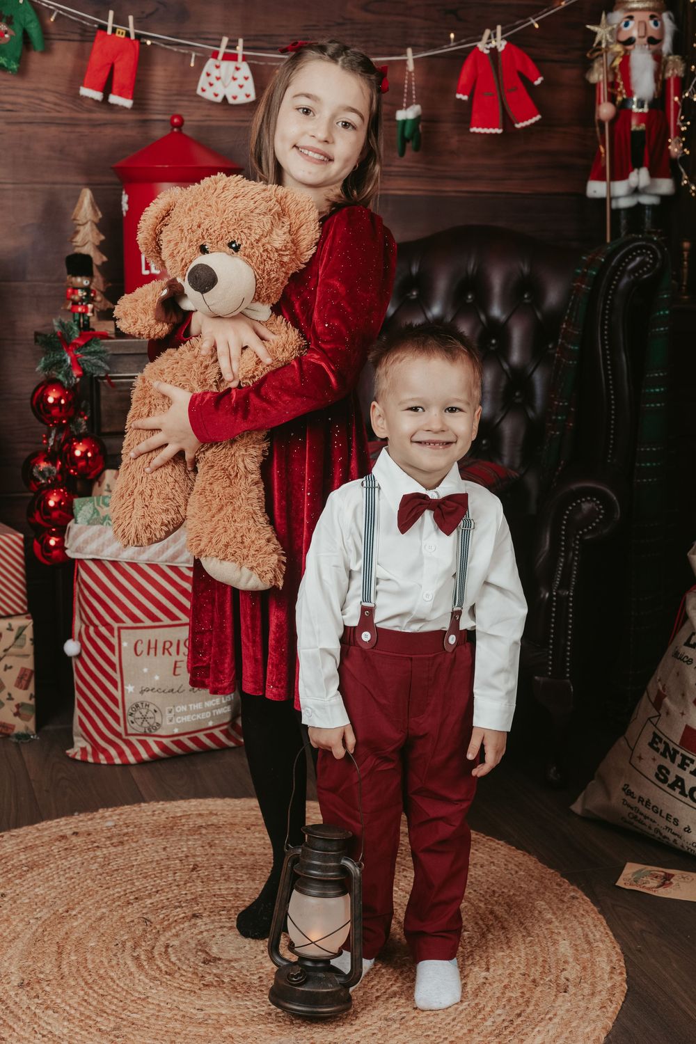 Children pose in festive red and white holiday outfits next to Christmas decorations and teddy bears on a round jute rug.