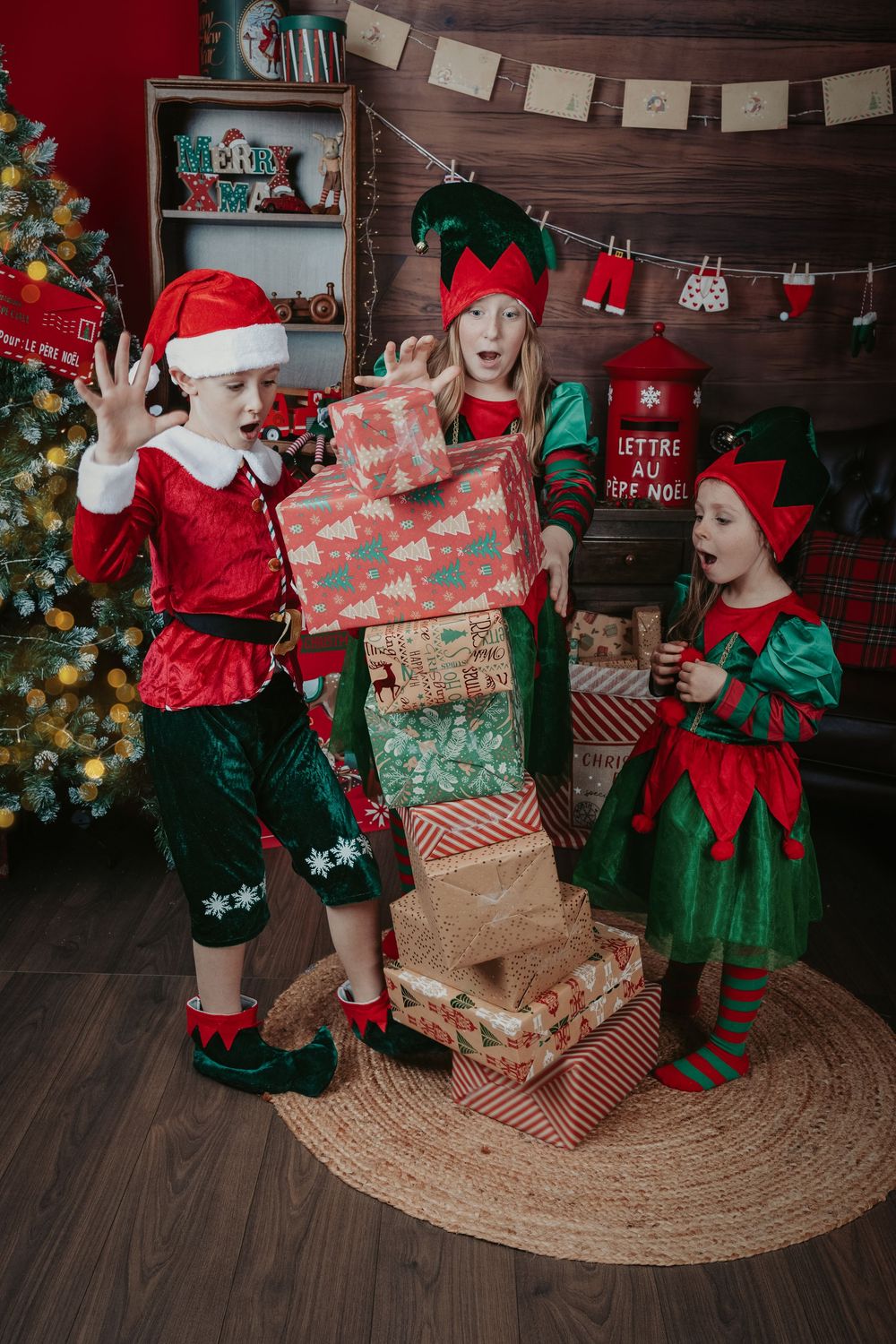 Two small figures in festive red and green elf costumes play with wrapped Christmas presents on a wooden floor.
