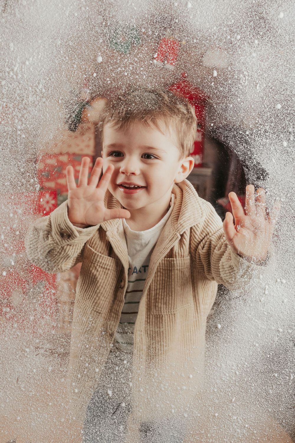 A joyful young child wearing a beige corduroy jacket presses hands against a frosted window with a festive red background.