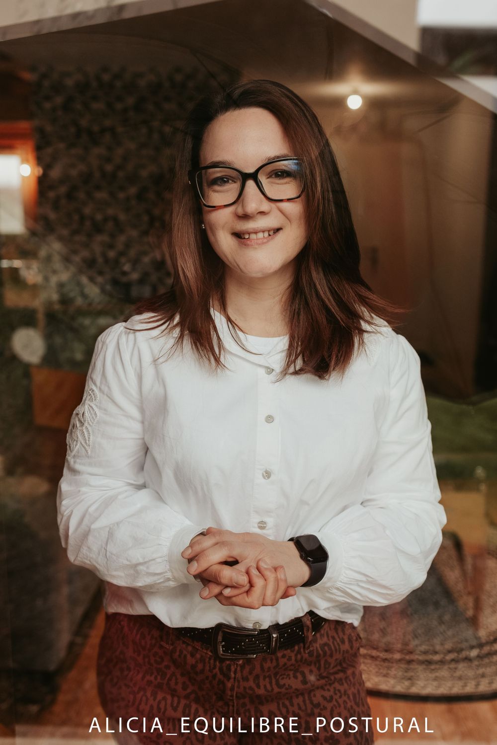 Person in white button-up top and glasses sits with clasped hands in warm indoor lighting.