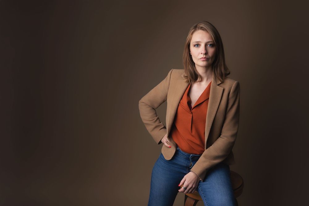 Woman in camel blazer and rust blouse poses confidently against a brown studio background.