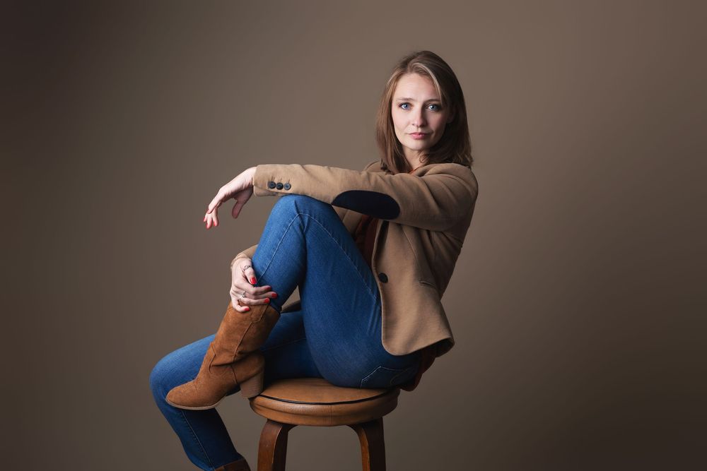 Woman in blue jeans, brown boots, and tan blazer sits confidently on a wooden stool against a gray background.