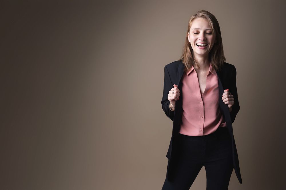 A cheerful businesswoman in a black blazer and pink blouse celebrates with fists raised against a neutral background.