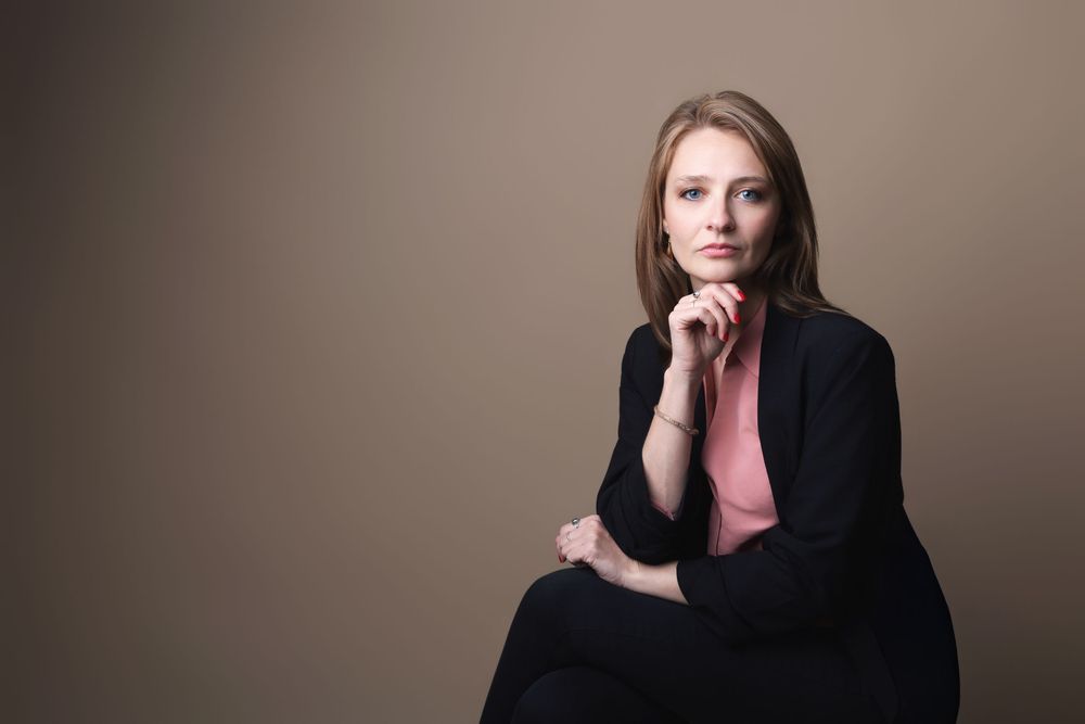 Professional woman in black blazer and pink top sitting confidently against neutral background.