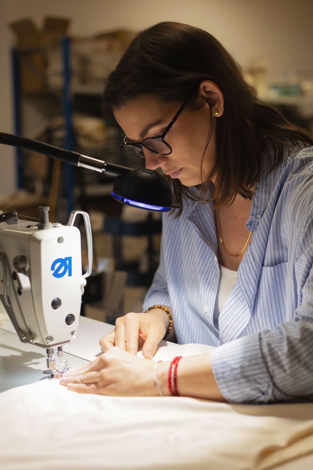 Woman with glasses sewing fabric at a machine under a blue lamp in a workshop.