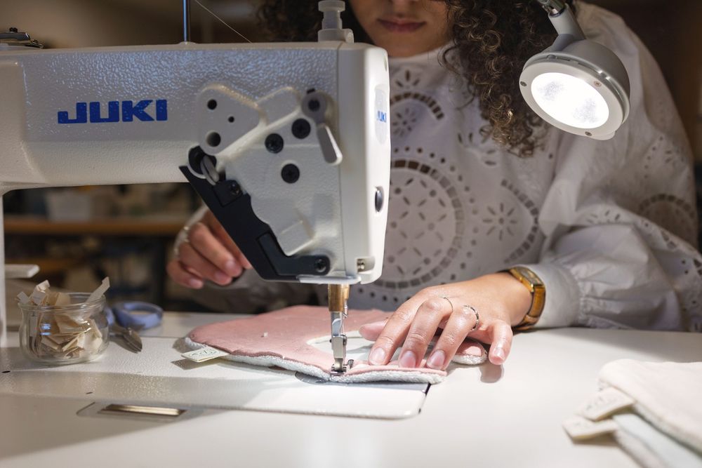 Woman sewing pink fabric on a Juki industrial sewing machine under bright lamp in a professional tailoring studio.