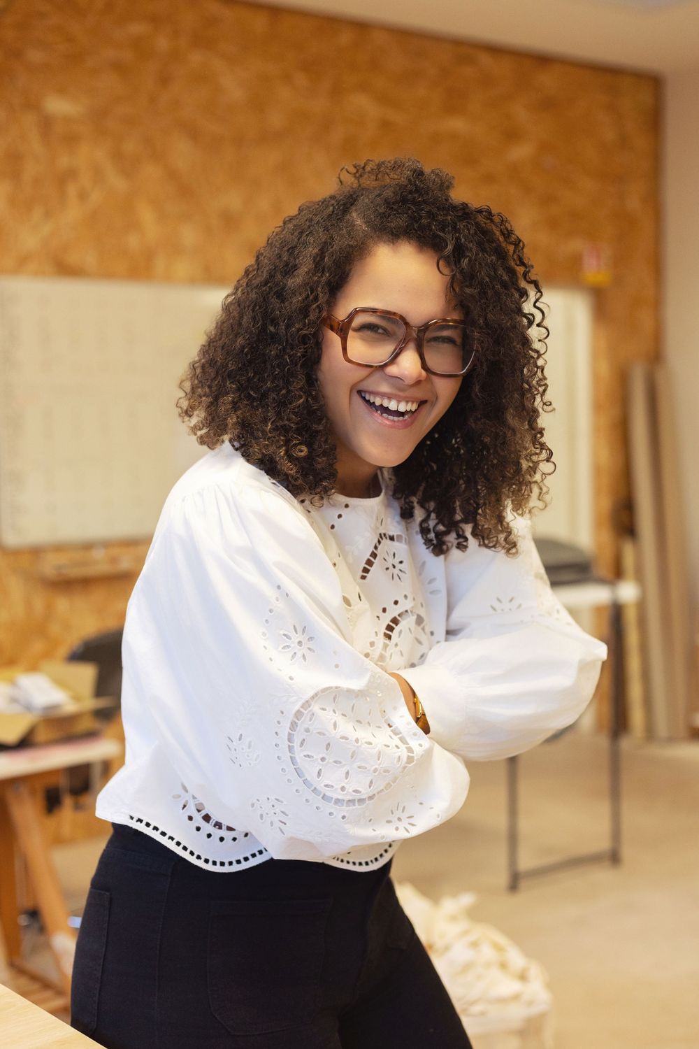 Smiling young woman with curly hair and glasses wearing a white blouse, arms crossed, standing in a workshop.