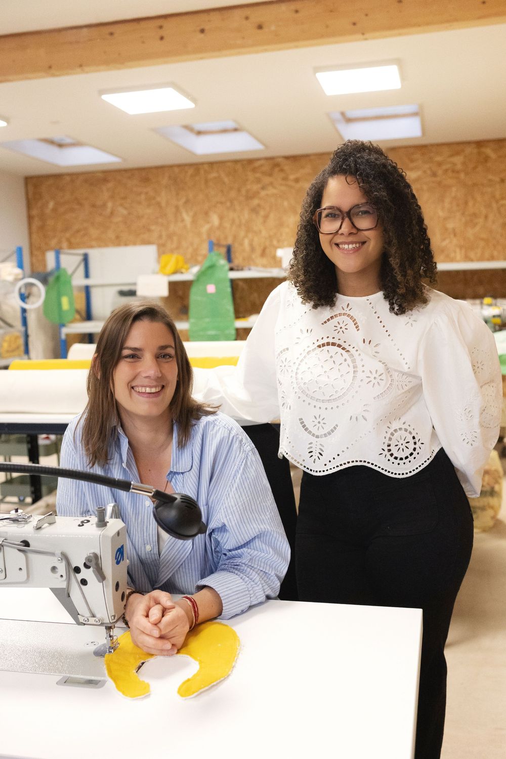 Two women smiling in a sewing workshop, one seated at a sewing machine, the other standing beside her.