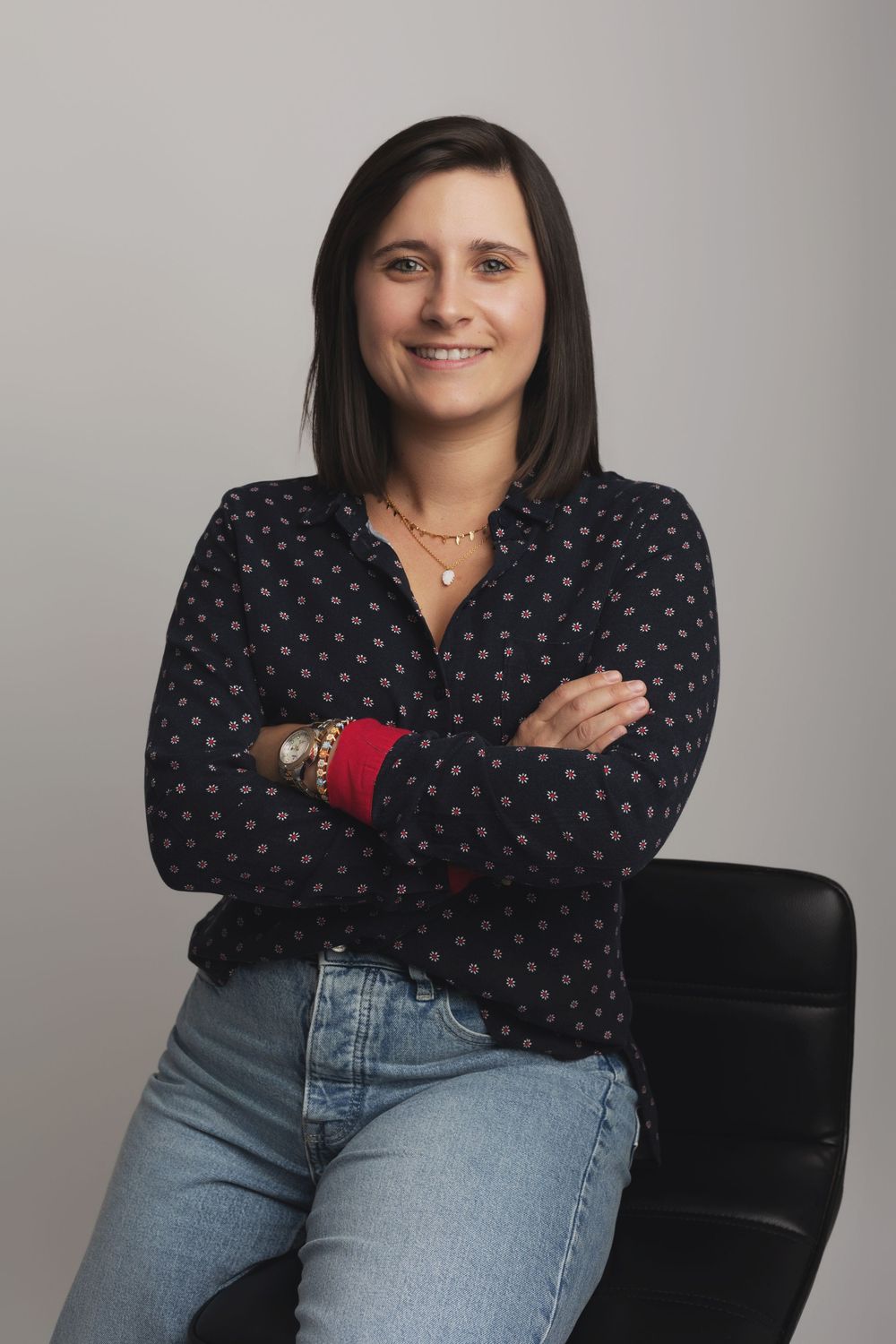 Woman in dark polka dot blouse and jeans sits cross-armed smiling in studio portrait.