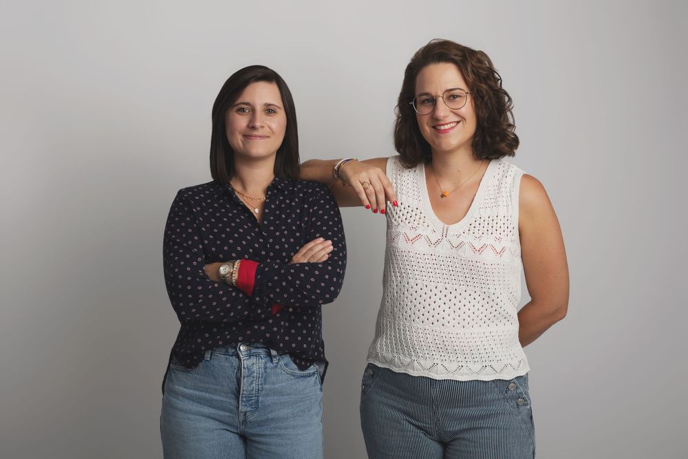 Two women smiling and posing together against a light gray background in casual attire.