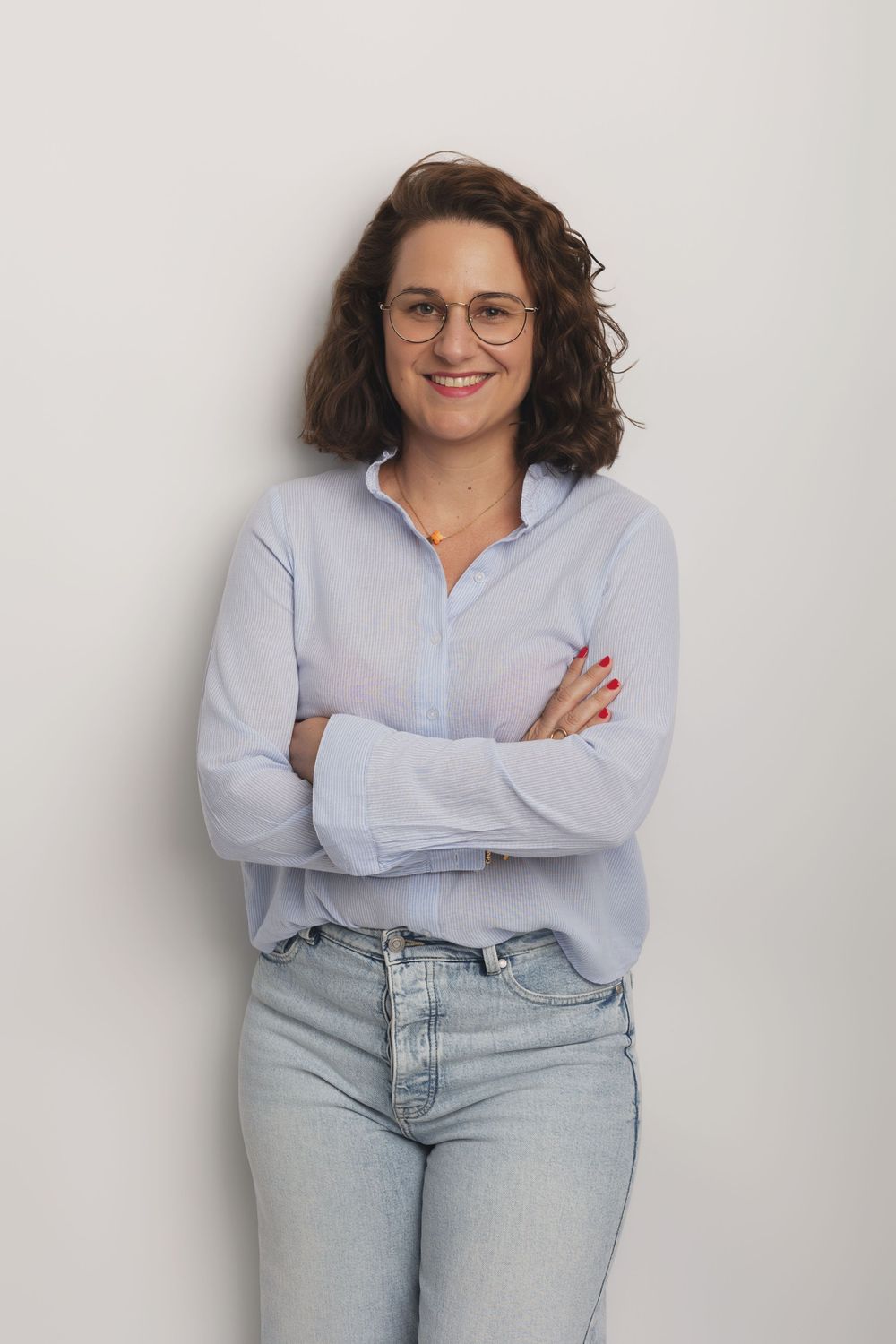 Smiling woman in light blue shirt and jeans with glasses poses arms crossed in studio.