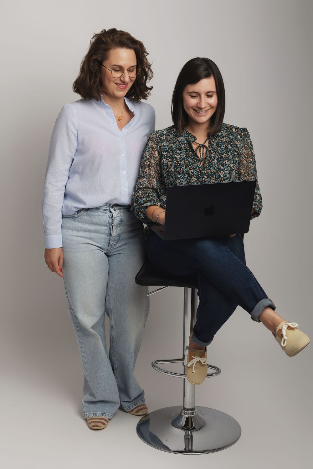 Two women smiling at a laptop, one seated on a bar stool, in a professional studio setting.