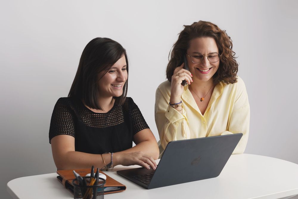 Two women smiling and collaborating on a laptop at a white desk in a bright office setting.