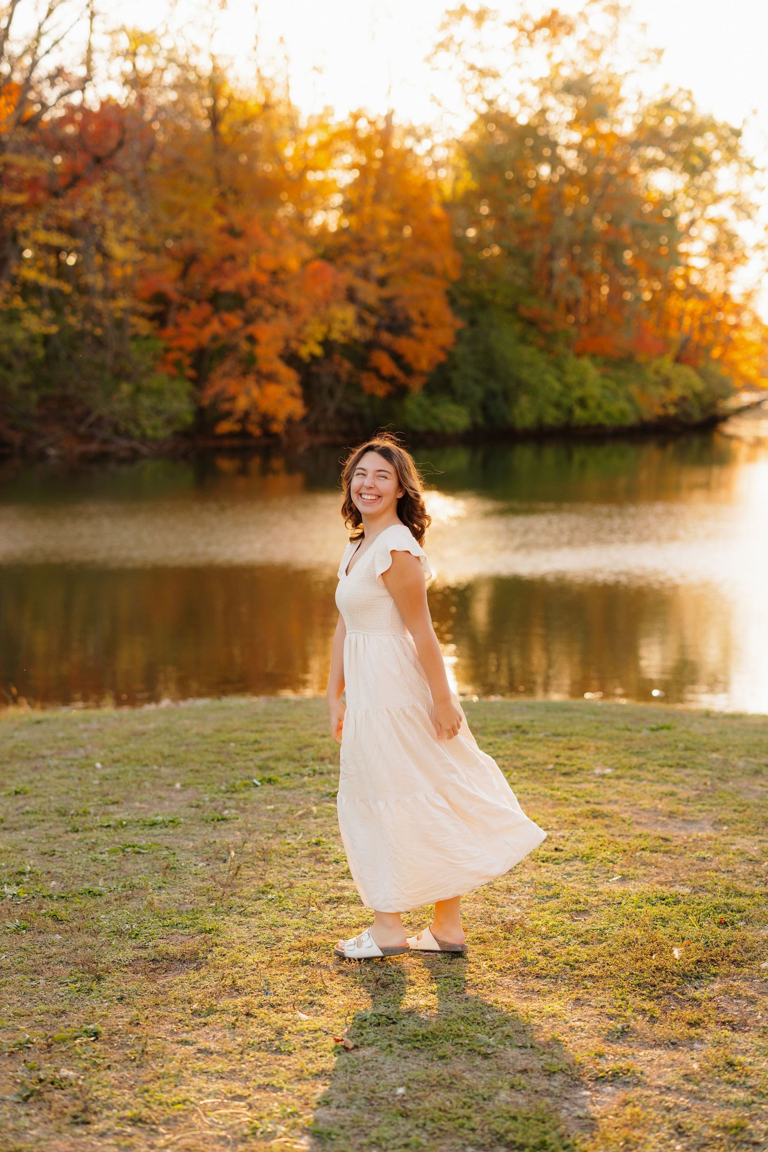 Someone in a flowing white dress twirls by an autumn lake with orange fall foliage reflecting in the calm water at sunset.