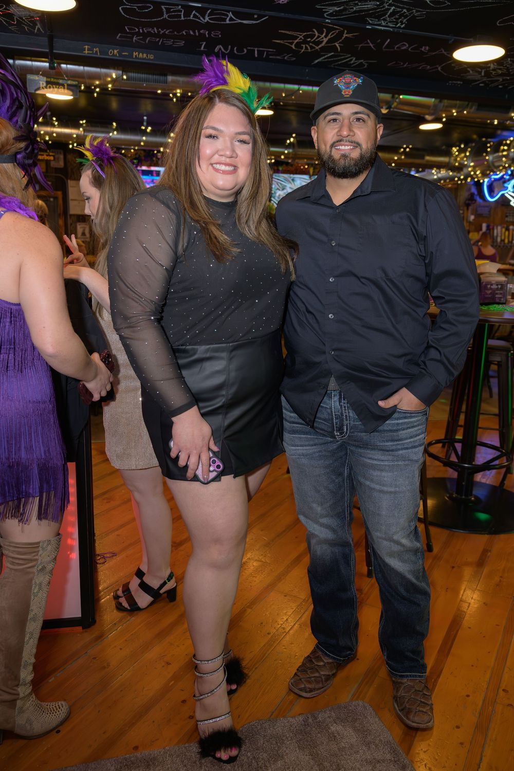 A couple poses together smiling at a Mardi Gras themed bar event, woman in black dress, man in dark shirt.