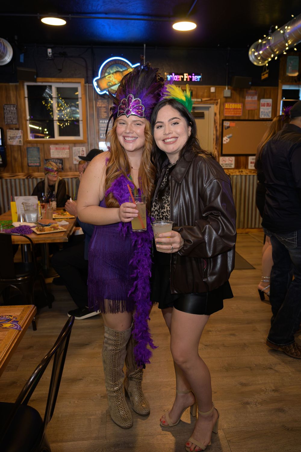 Two women in Mardi Gras costumes pose with drinks at a festive bar celebration.