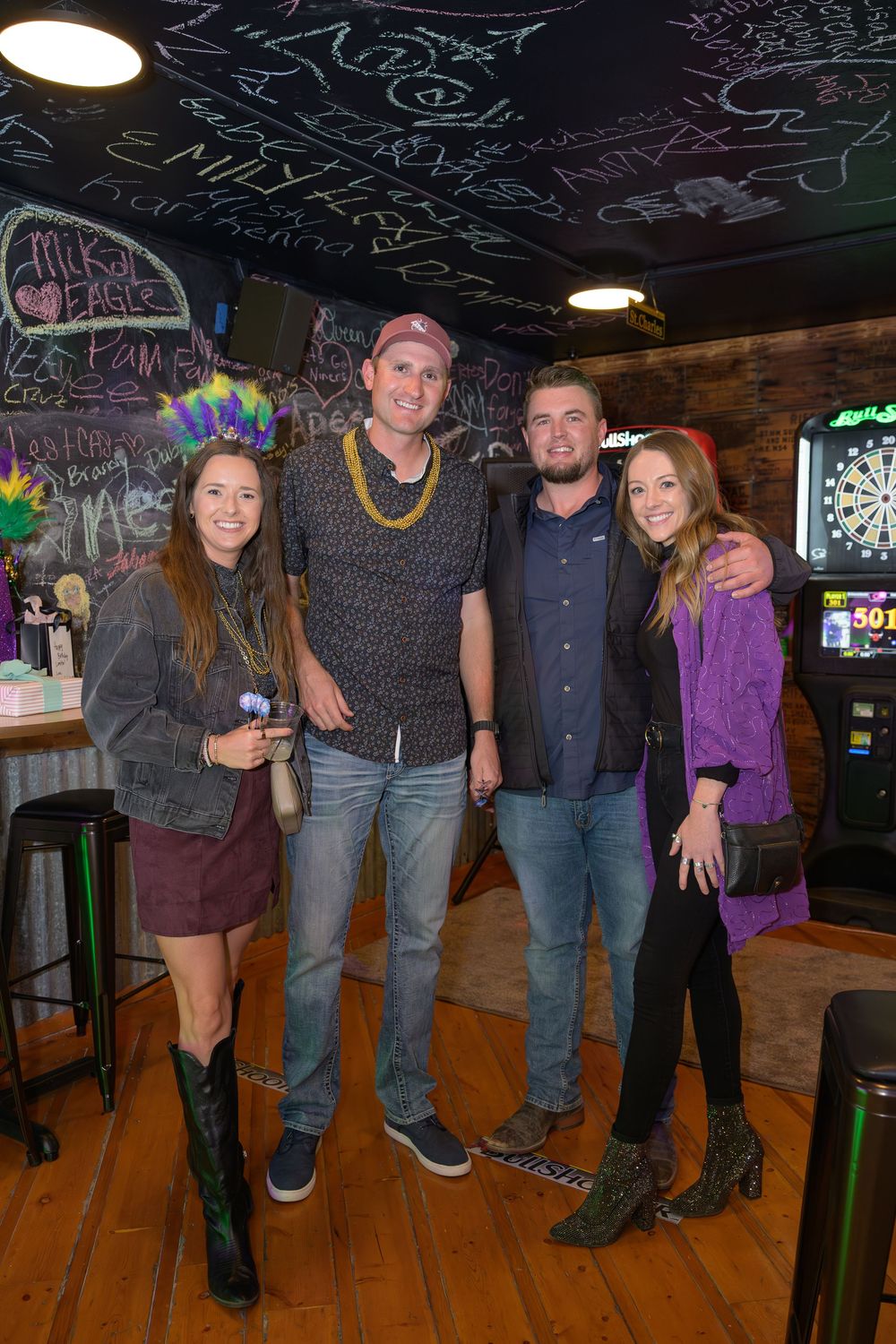 Four friends pose together at a Mardi Gras party in a bar with chalkboard walls and a dartboard.