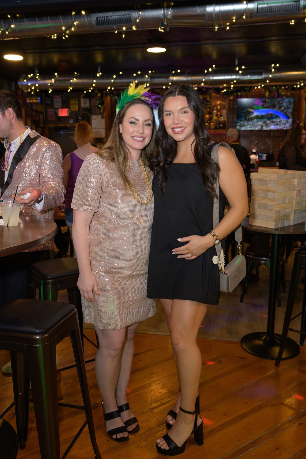 Two women pose together at a Mardi Gras party, one in a gold sequin dress with feather headband, other in black mini dress.