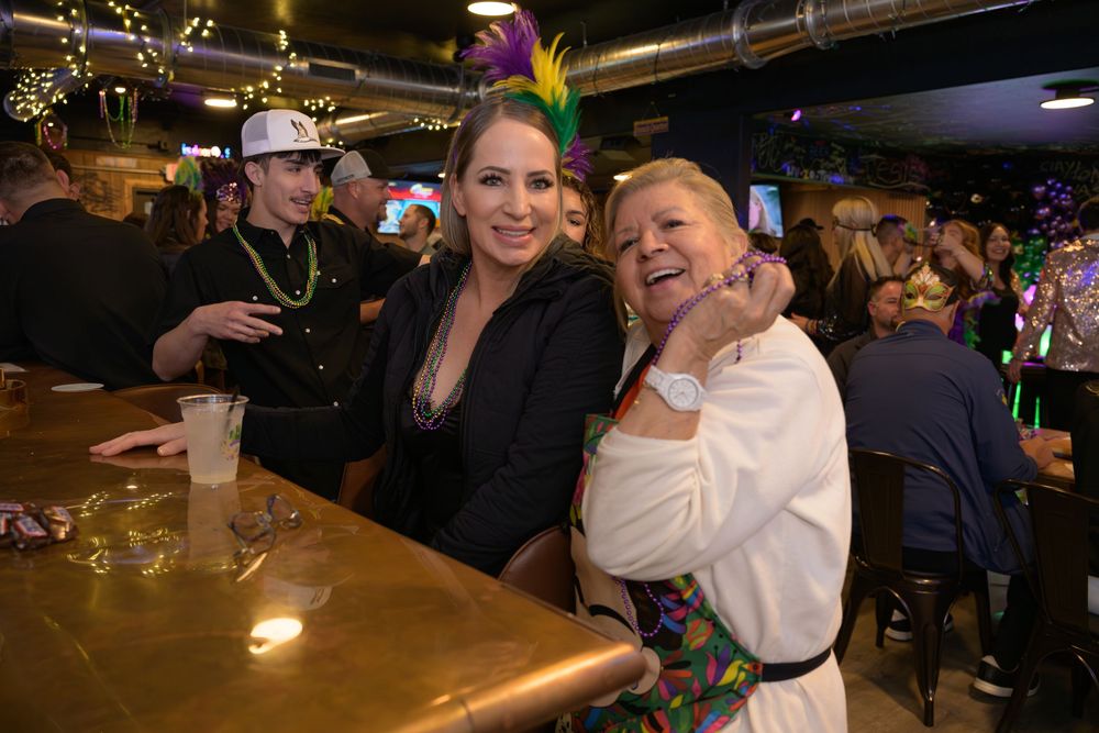 Two women pose together at a Mardi Gras bar celebration, wearing festive beads and feathered accessories.