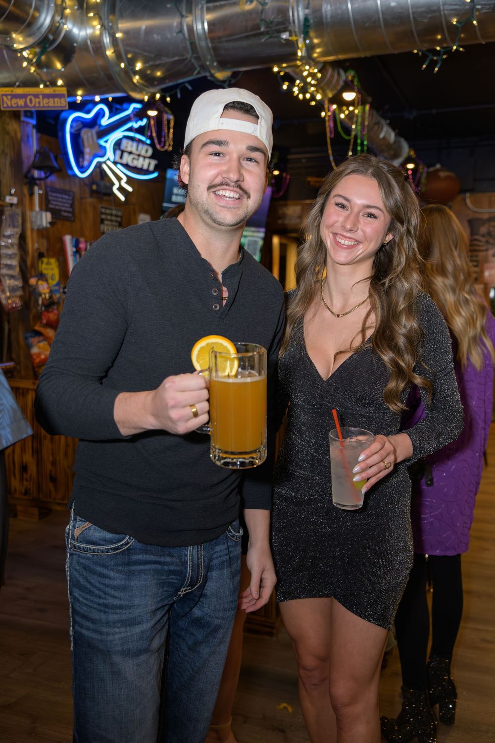 Smiling couple poses with drinks at a lively New Orleans-themed bar celebration.