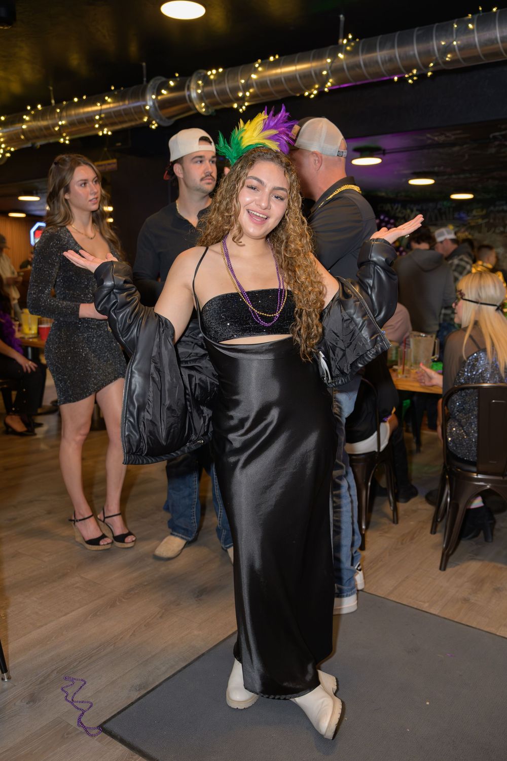 Woman in black satin dress with Mardi Gras feather headpiece poses at a festive indoor party.