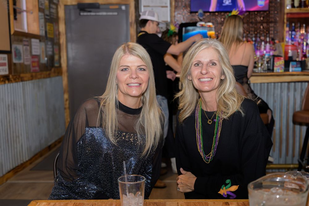Two smiling women pose together at a bar, one in a sparkly top and the other wearing Mardi Gras beads.