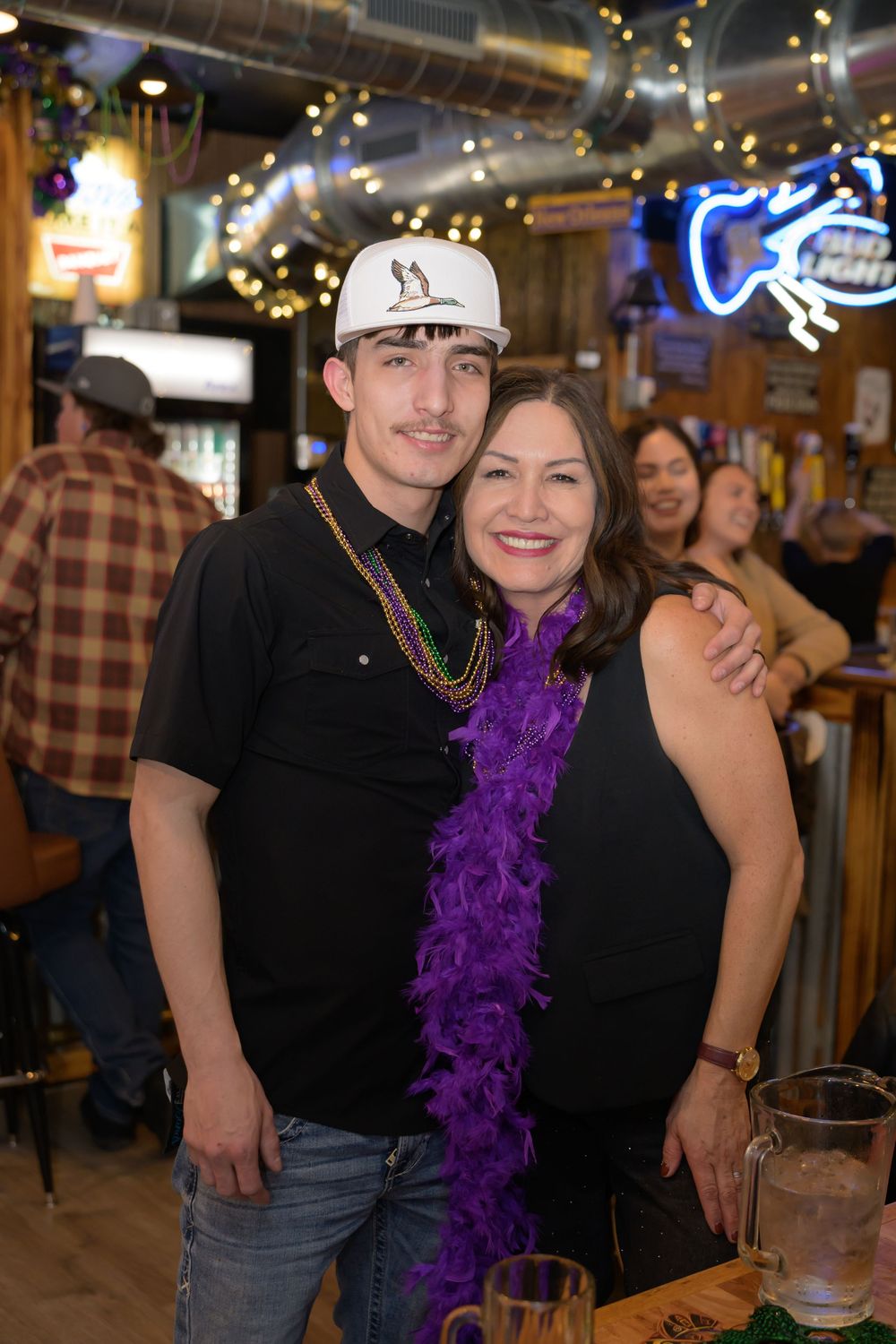 A young man and woman smile together wearing Mardi Gras beads and purple boa at a bar.