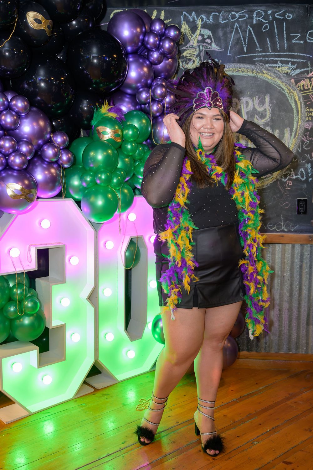 Woman in Mardi Gras feather boa and headdress posing at a 30th birthday party with purple and green balloons.
