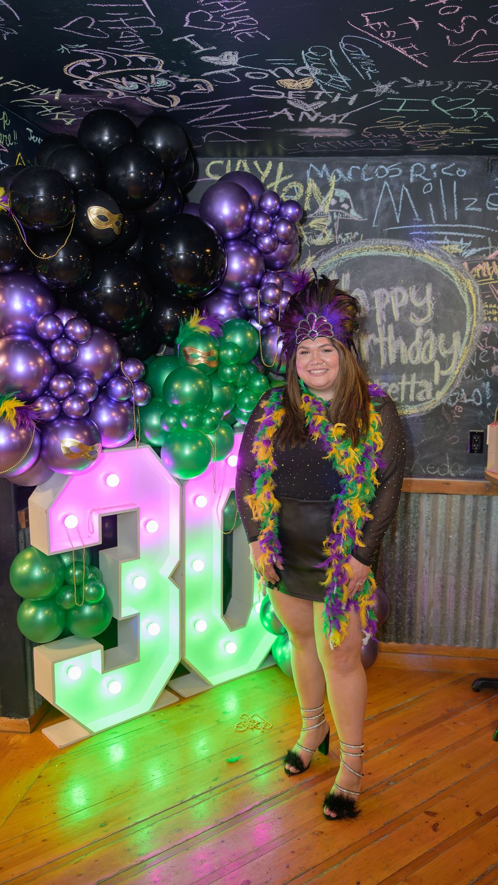Woman in Mardi Gras attire poses by illuminated 30th birthday marquee letters with purple and green balloons.