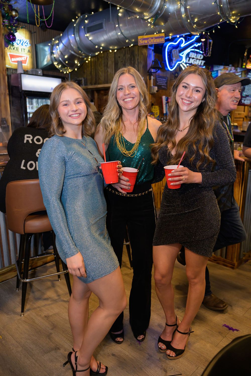Three women pose together at a bar holding red cups, dressed in elegant party outfits.