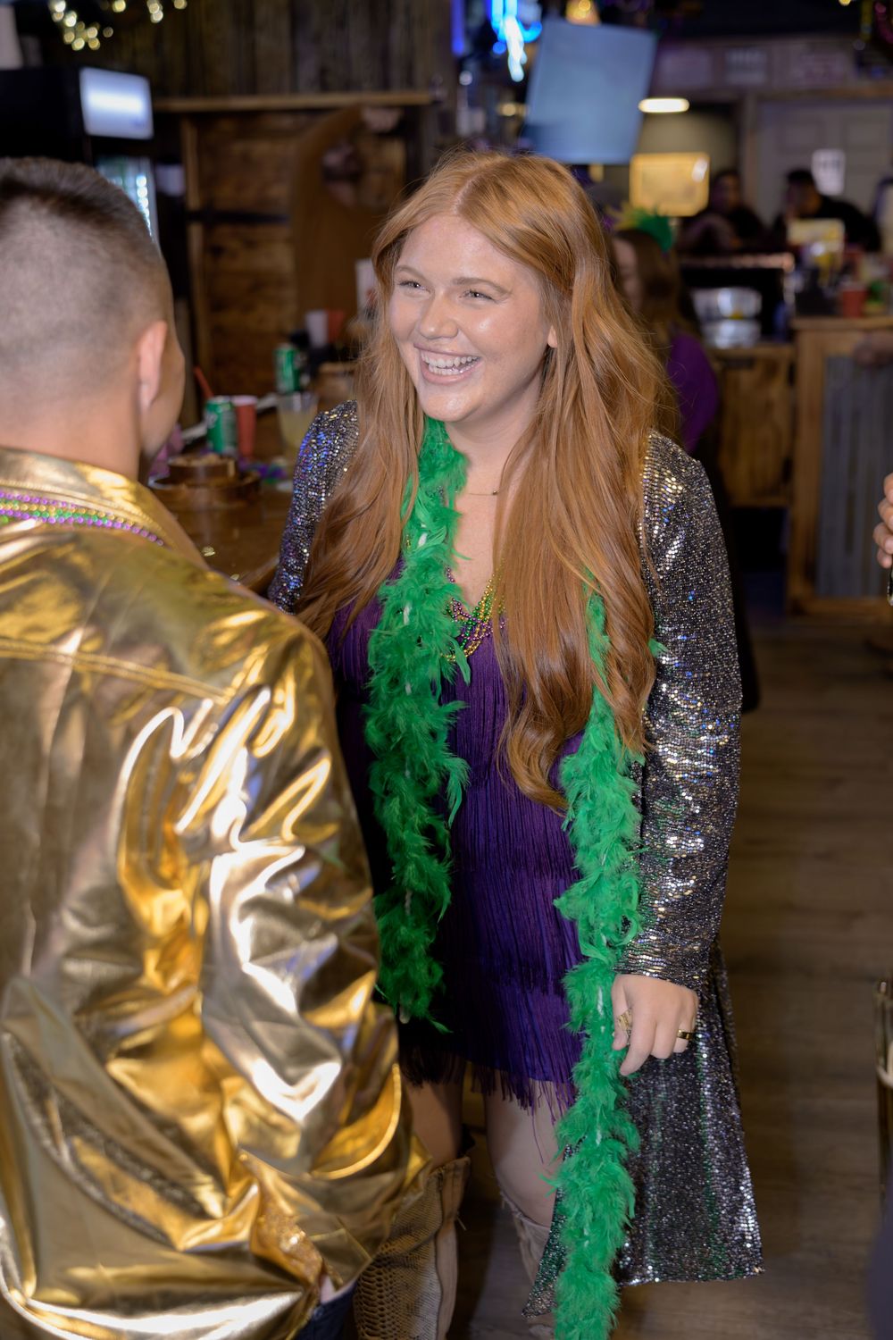 A smiling redhead woman wearing a green feather boa and purple dress talks with a man in a gold jacket at a Mardi Gras party.