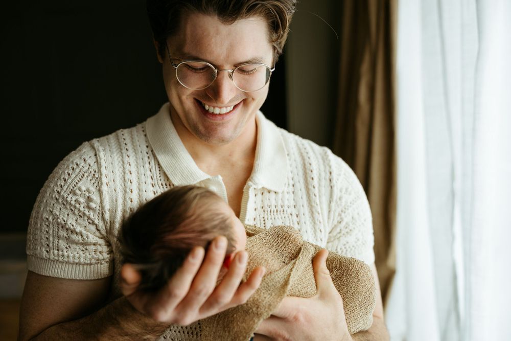 Caregiver in white knit sweater holds and gazes lovingly at an infant near a window with soft lighting.
