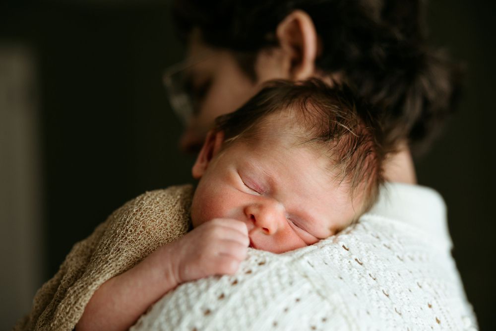 A sleeping newborn baby with closed eyes rests peacefully against someone's shoulder wrapped in a knitted blanket.