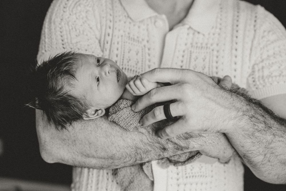 A black and white photograph shows tender hands cradling a small infant in delicate white fabric.