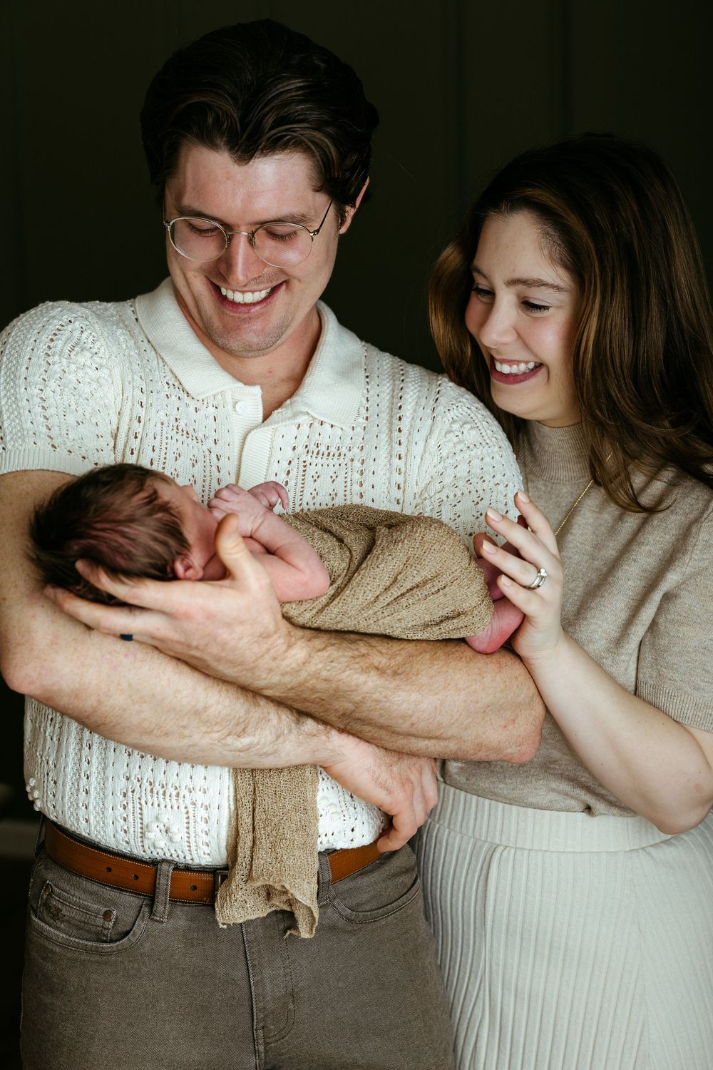 Two new parents sharing a joyful moment as they cradle their newborn baby wrapped in a beige blanket.
