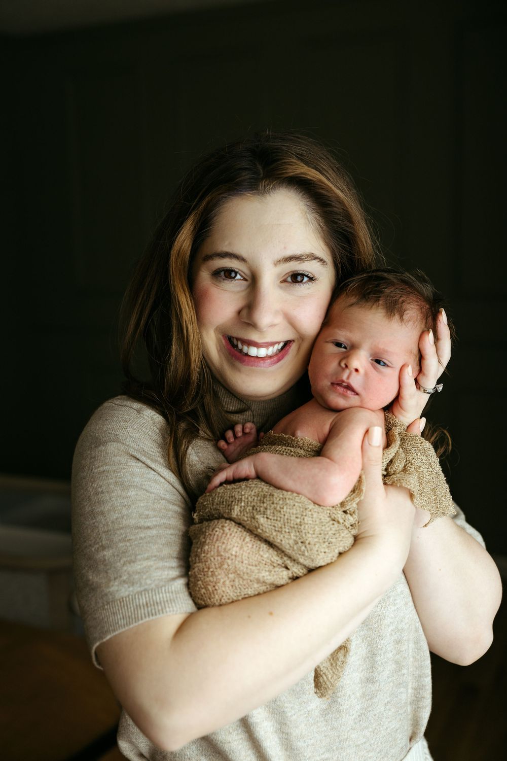 A heartwarming portrait shows a mother in a gray shirt holding her newborn baby wrapped in a beige towel against a dark background.