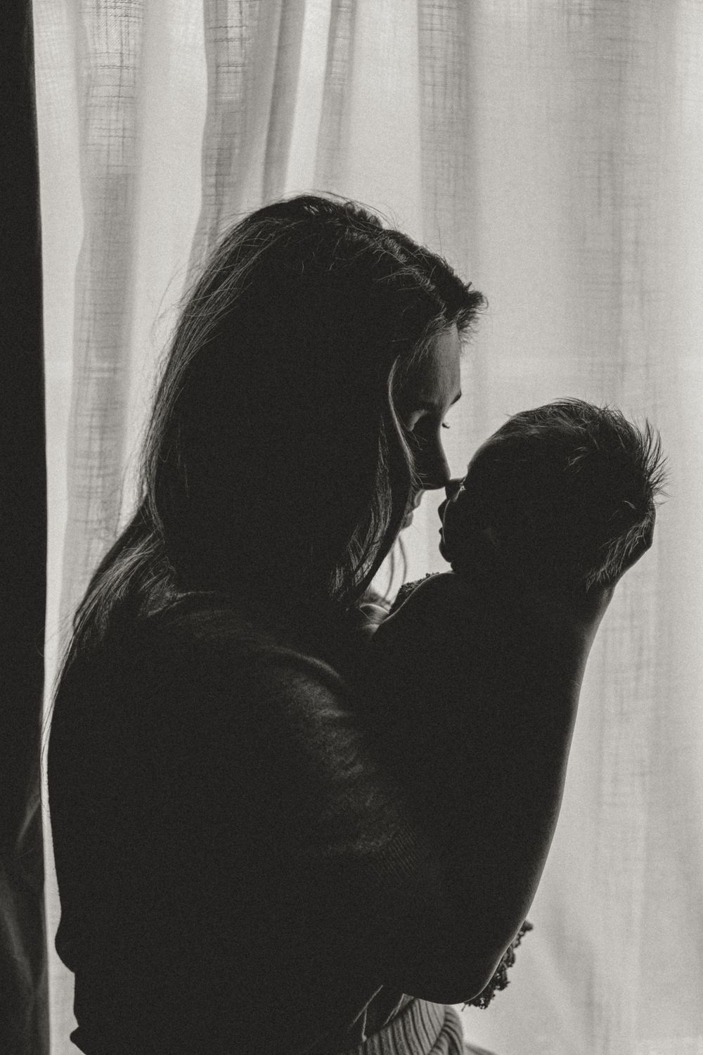 A silhouetted profile of a parent holding a baby against soft window light creating a tender black and white portrait.