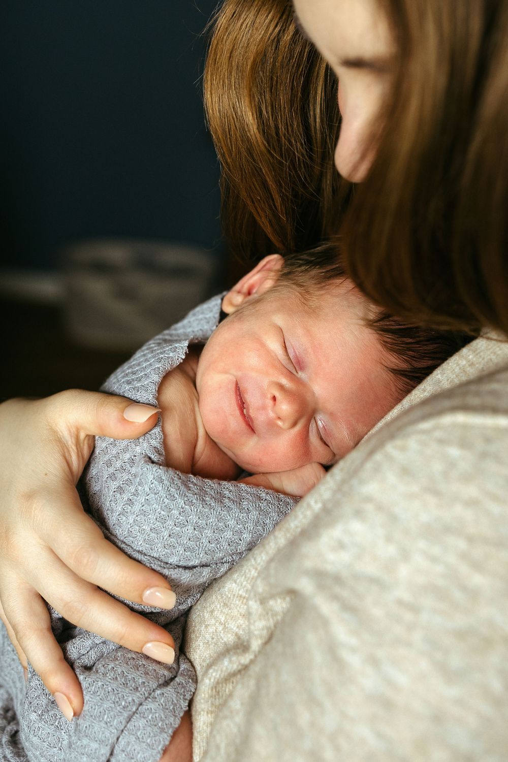 A newborn baby in a light gray onesie sleeps peacefully while being held in loving arms.